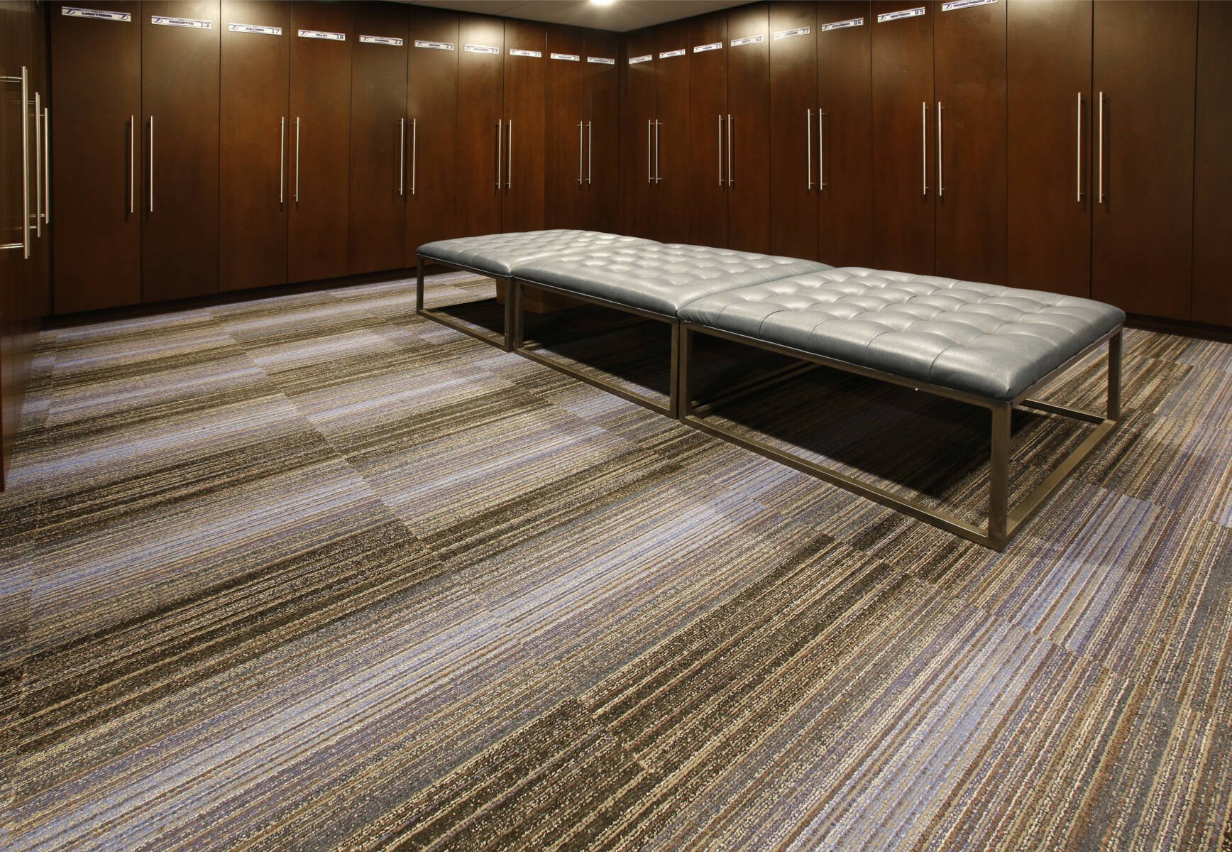 Locker room with brown and white striped carpet tile, cherry wood cabinets and white pincushion benches. This image demonstrates this company's excellence in multi-purpose sports flooring installations.